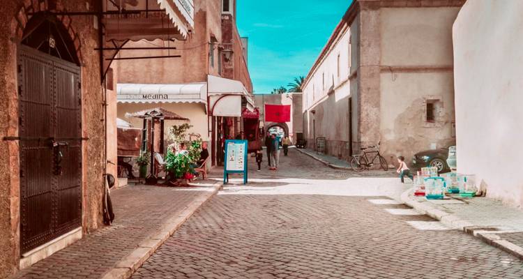 Calle estrecha de la medina con adoquines, llena de tienditas, toldos y gente local bajo un cielo azul vibrante.