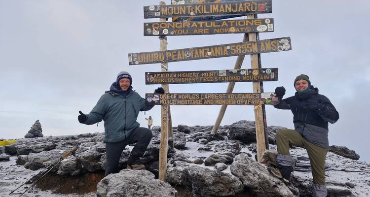 Two triumphant trekkers pose by the Uhuru Peak sign atop snow-dusted Mount Kilimanjaro.