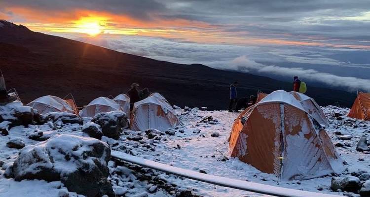 Snow-dusted orange tents dot a high-altitude campsite at sunrise above a sea of clouds on Kilimanjaro.