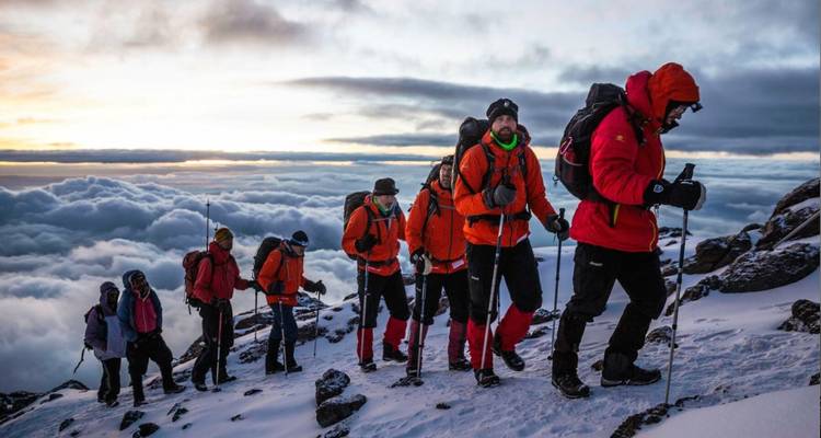 A rope of climbers in bright jackets ascend a snowy ridge above a blanket of clouds at dawn.