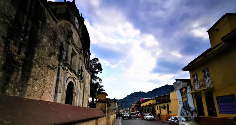 Colonial-era church and colourful street with cars and locals, backed by mountains under dramatic clouds.