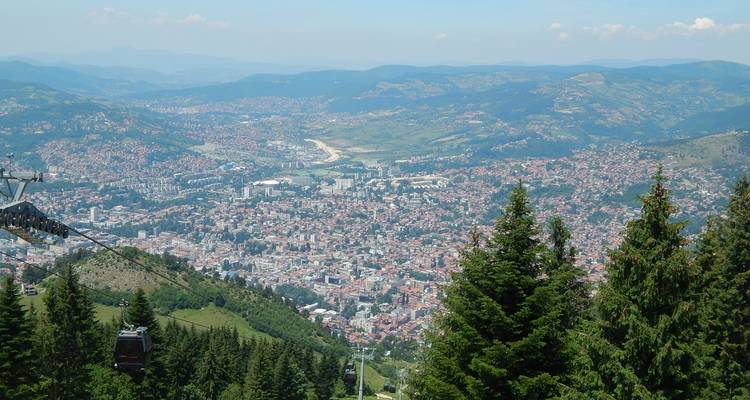 Uitgestrekt luchtfoto van de stad Sarajevo omlijst door naaldbos en kabelbaan.