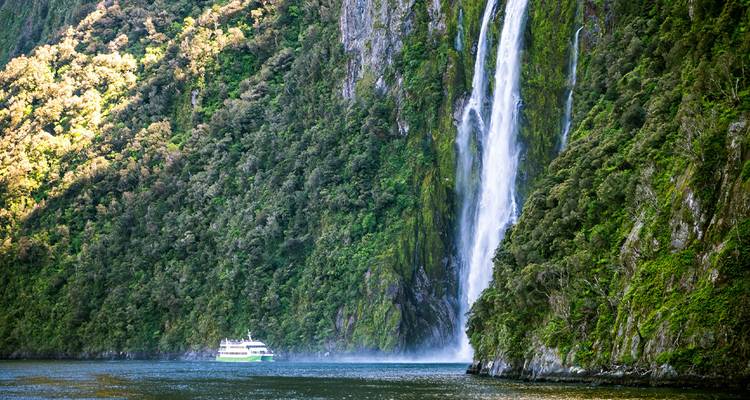 Tall waterfall dropping down lush vertical cliffs into Milford Sound with tour boat below for scale.