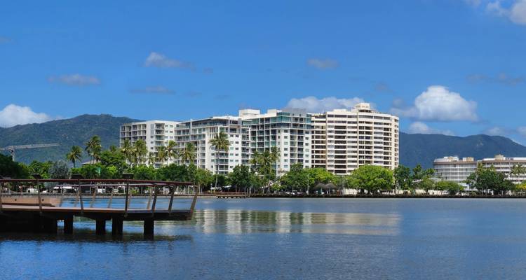 Modern waterfront apartments in Cairns reflected on calm inlet waters with forested hills behind.