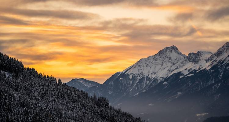 Montañas cubiertas de nieve al atardecer con cielo naranja vívido sobre una cresta boscosa oscura