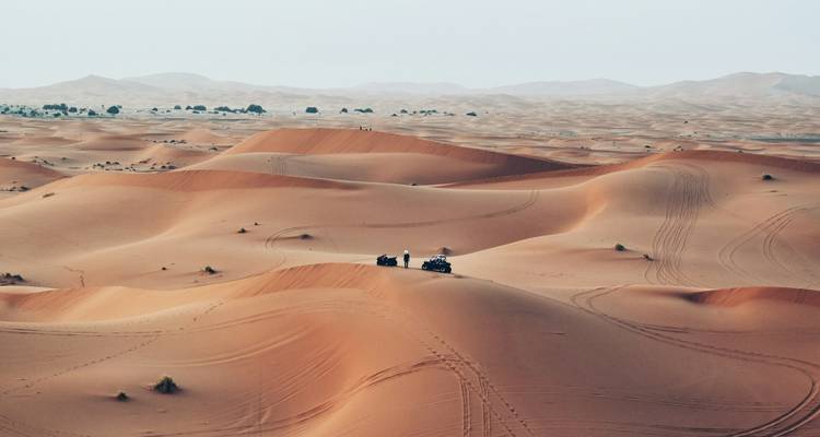 Vastas dunas de arena naranja se extienden hasta el horizonte mientras dos viajeros en cuatrimotos hacen una pausa en una cresta bajo un cielo pálido.