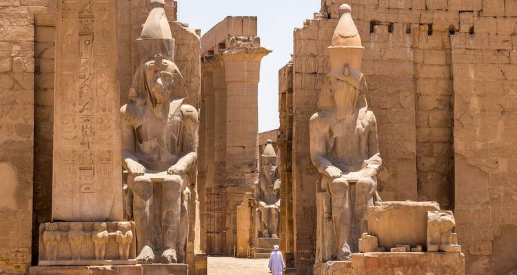 Gigantische sitzende Statuen und papyrusförmige Säulen säumen eine Allee im Luxor-Tempel, während ein einzelner Besucher zusieht.