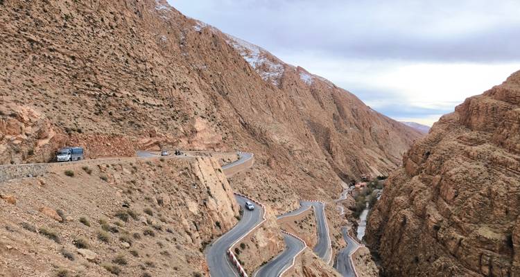 Serpentine mountain road carved into steep reddish cliffs with vehicles climbing toward snow-tipped peaks.
