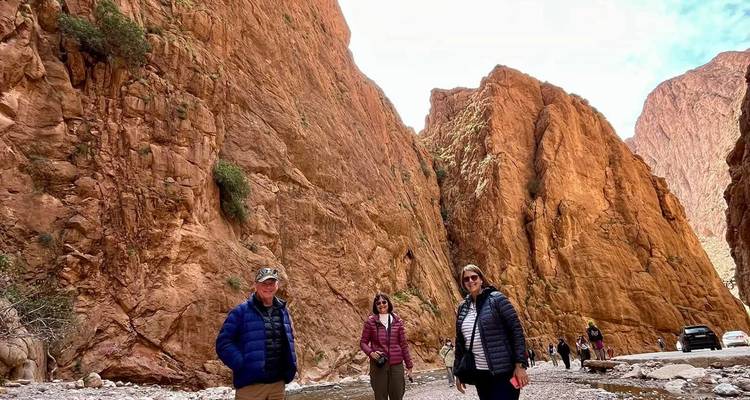 Visitors standing beside towering orange canyon walls with a shallow stream at their feet.