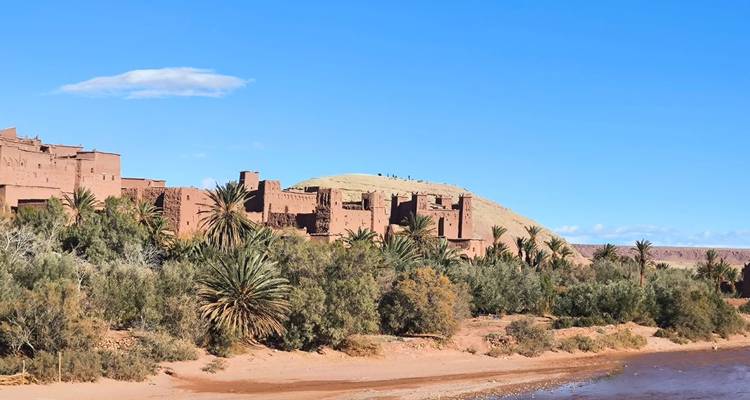 Adobe kasbah of Ait Benhaddou rising above palm trees and a dry riverbed in bright sunshine.
