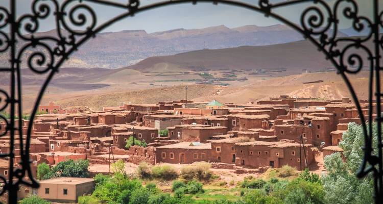 Rustic adobe village and green valley framed by decorative wrought-iron window arch.