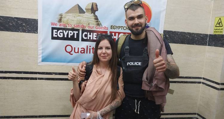 Zwei Reisende zeigen Daumen hoch vor einem Werbebanner für Ägypten-Touren in einem Innenraum.