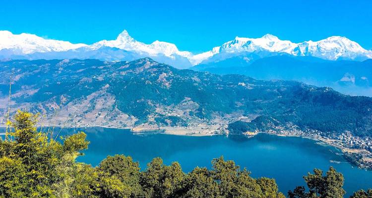 Panoramablick auf einen großen blauen See, umgeben von üppigen Hügeln mit der schneebedeckten Annapurna-Bergkette, die sich im Hintergrund unter einem klaren blauen Himmel erhebt