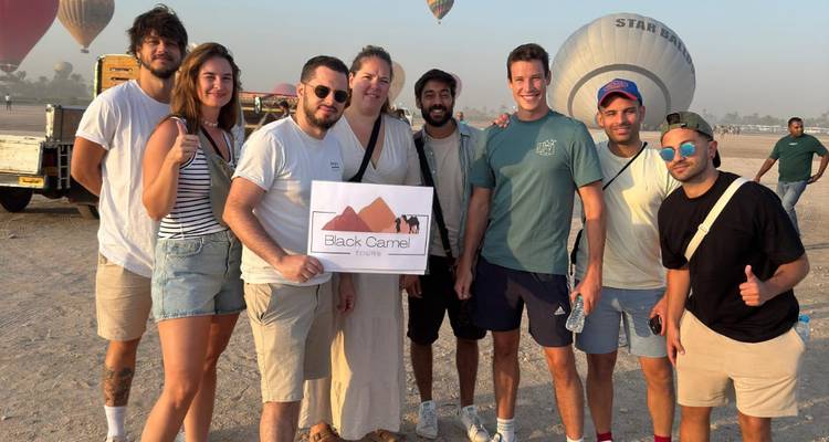 Smiling tour group holding a 'Black Camel Tours' sign on sandy field with hot air balloons behind.