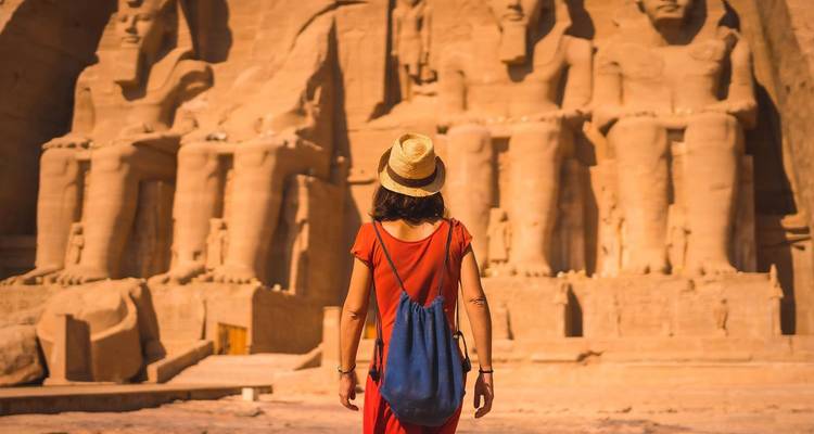 Back view of a woman in a hat approaching the colossal statues of Abu Simbel under warm light.