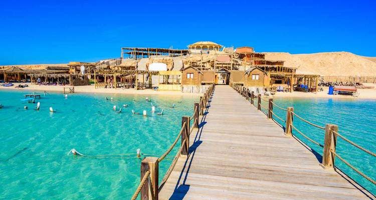 Wooden pier leads over crystal-clear turquoise water to rustic beachfront huts on a sandy island.