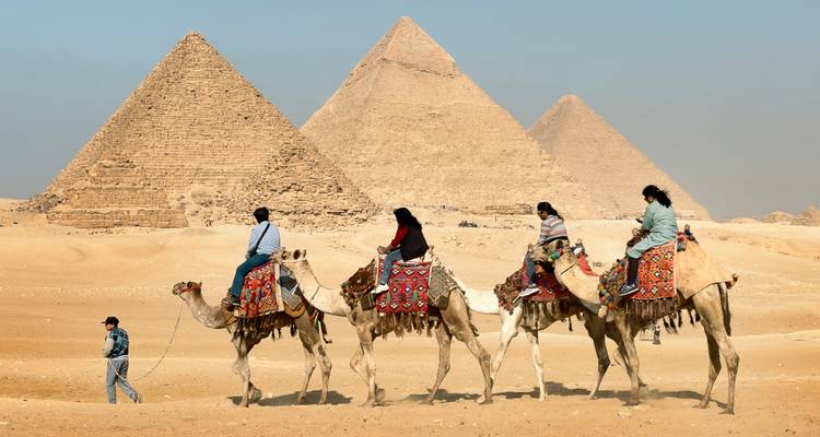 Line of tourists riding camels in front of the Great Pyramids across sandy desert.