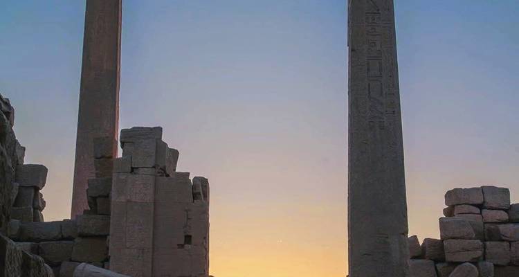 Two ancient obelisks and crumbling stone walls silhouetted against a pastel sunset.