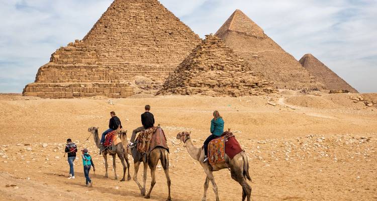 Travellers riding camels across the sands with the Pyramids towering behind.