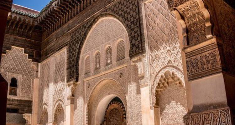 Patio bañado por el sol de una madrasa marroquí histórica con arcos de yeso tallado y aleros de madera.