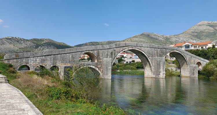 Puente de arco de piedra histórico que atraviesa un río turquesa cristalino con montañas y pueblos de fondo.