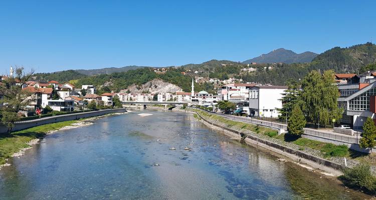 A calm river winds through a Bosnian town backed by rolling green hills and a clear blue sky.