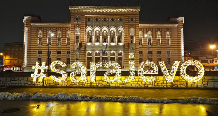 Illuminated #saraJevo sign sparkles in front of the ornate Sarajevo City Hall at night, reflecting on wet pavement.