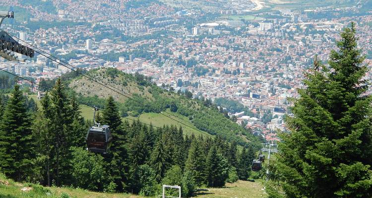 Cable cars glide above dense pine forest toward a panoramic view of Sarajevo spread below.