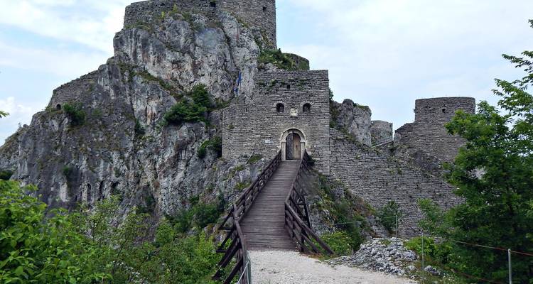 A dramatic medieval stone fortress perched on a rocky cliff is reached by a narrow wooden bridge.