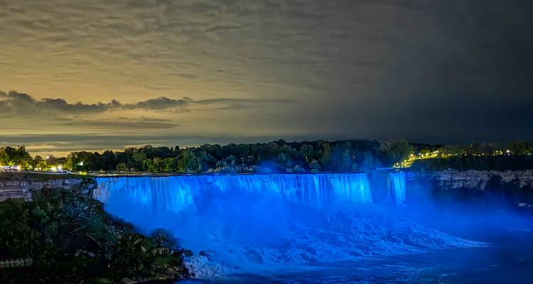 Niagara Falls illuminated in blue at night with mist rising against a dramatic sky.