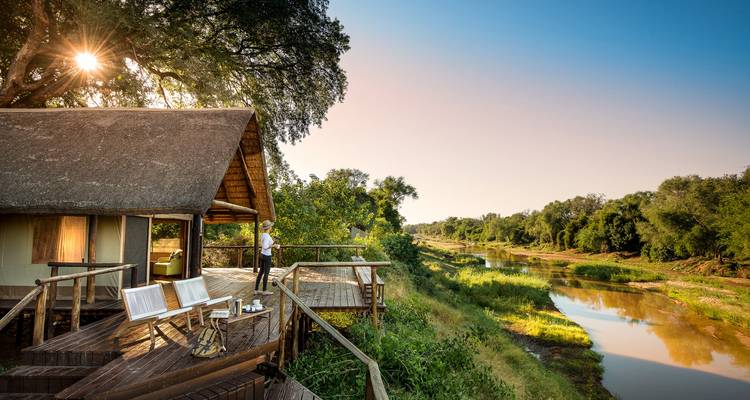 Wooden deck of a thatched safari lodge with a guest enjoying a sunrise view over the river
