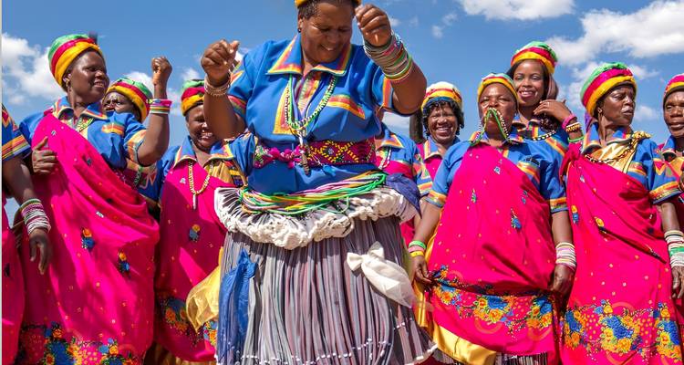 Vibrant group of women in traditional South African attire dancing joyfully beneath a sunny sky.