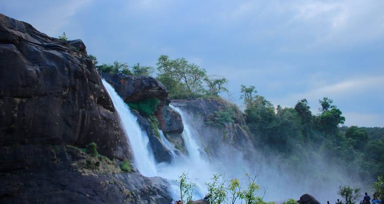 Twin torrents of a large waterfall cascade down a jungle-clad cliff, mist rising into a cloudy sky.