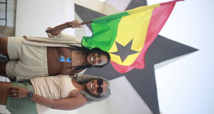 Two smiling women holding the Ghanaian flag in front of the Black Star Monument in Accra