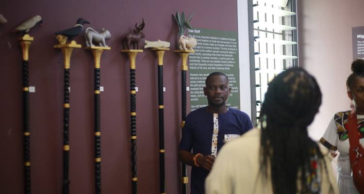 Museum guide explaining a display of ornately carved ceremonial staffs on a maroon wall