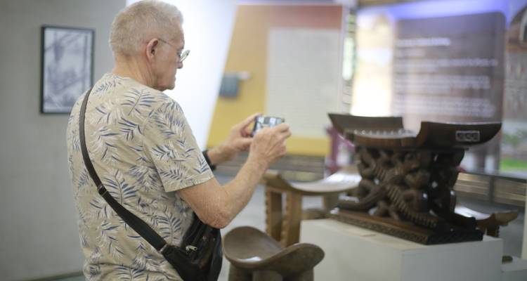 Senior tourist photographing a wooden royal stool exhibit in a Ghanaian museum.