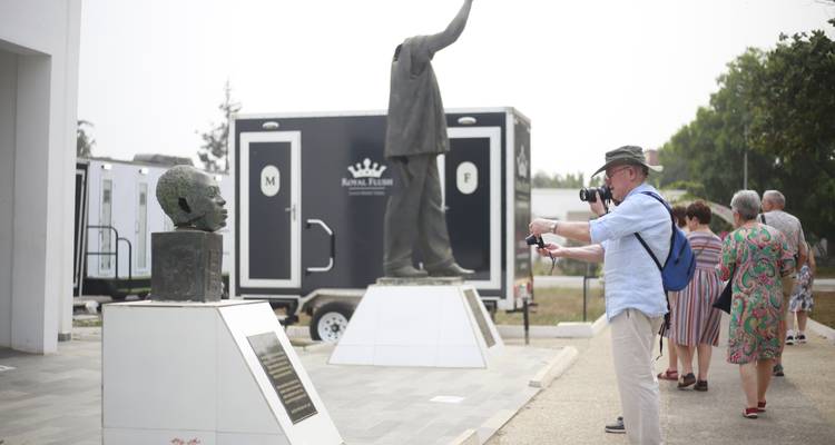Tourist photographing a bust and statue outside a memorial complex while companions walk ahead.