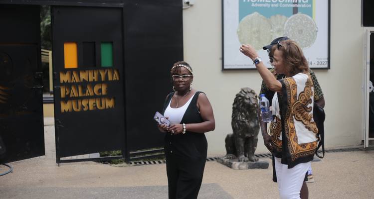 Tourists chatting outside the entrance gate of Manhyia Palace Museum featuring Ghanaian emblem.
