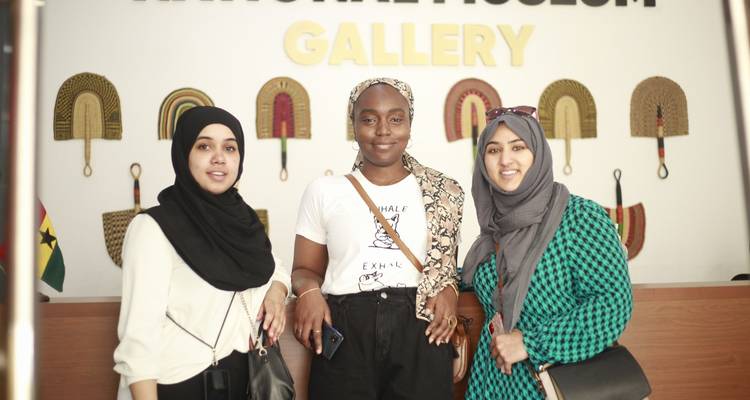 Friends smile for a photo inside the National Museum Gallery with decorative fans on the wall.