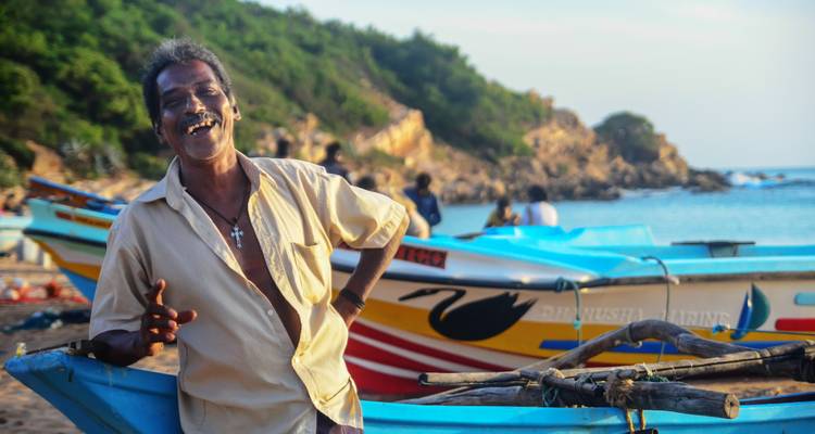 Un pêcheur local souriant pose à côté de bateaux aux couleurs vives sur une côte rocheuse du Sri Lanka.