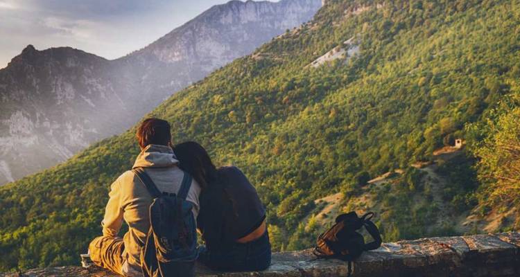 Young couple sitting on a stone ledge enjoying a tranquil view of forested mountains in soft evening light.