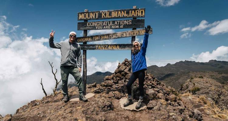Deux randonneurs triomphants lèvent les bras au panneau du sommet du mont Kilimandjaro avec des nuages en contrebas.