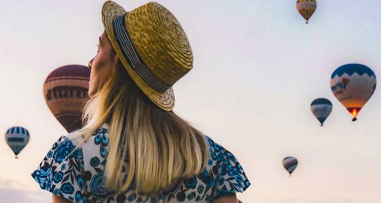 Woman in straw hat gazing at colorful hot-air balloons floating over Cappadocia’s sunrise sky.