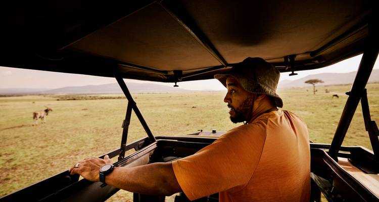 Traveller stands inside an open-top safari vehicle looking out over vast grassy plains.