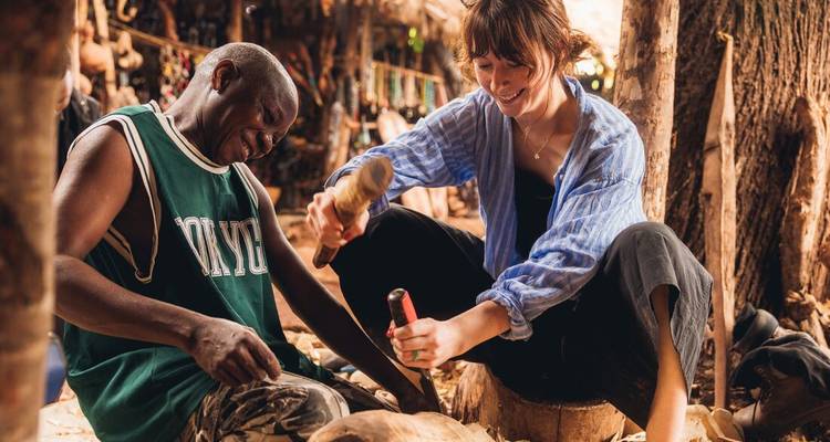 Local artisan teaches a traveller traditional wood carving inside a rustic workshop.