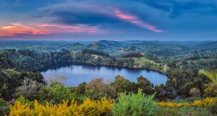 Vibrant sunset illuminates a circular crater lake surrounded by rolling green hills and forests.