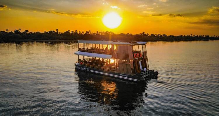 A double-deck river cruiser drifts across calm water beneath a fiery sunset sky.