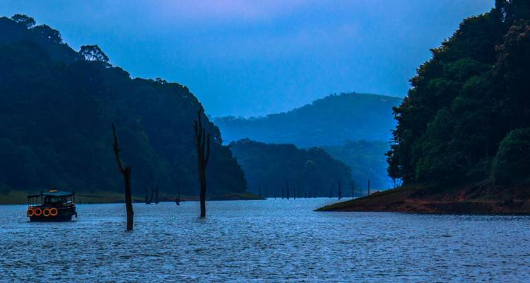 Misty blue hills frame Periyar Lake at dusk with a lone boat drifting near skeletal tree trunks.