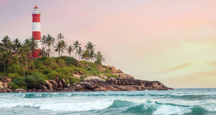 The red-and-white striped lighthouse rises above palm-covered rocks beside turquoise sea at Kovalam.