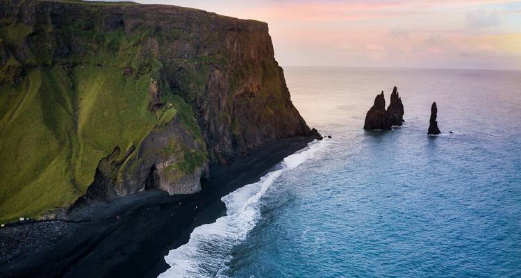 Vue aérienne de la plage de sable noir de Reynisfjara et des formations rocheuses de Reynisdrangar à l'aube aux teintes pastel.